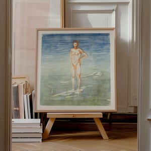May include: A framed print of a nude figure in water, with blue and green tones. The artwork is displayed on a small wooden easel. Other framed art and books are visible in the background. The frame is a light wood colour.