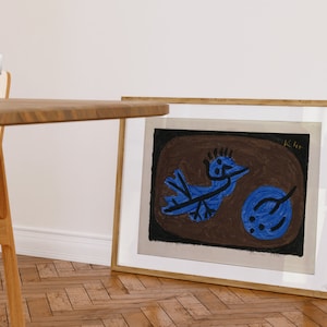 Op de afbeelding: Een ingelijste print van een blauwe vogel met zwarte contouren op een bruine achtergrond. De handtekening van de kunstenaar "Klee" staat in de rechterbovenhoek van de print.