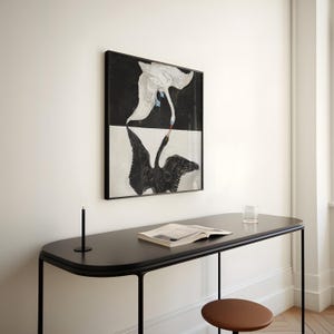 May include: A black and white framed print of two swans, one white and one black, with their necks intertwined. The print is hanging on a white wall above a black desk with a book and a glass on it.