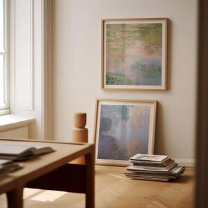 May include: Two framed art prints with light wood frames hang on a beige wall. The top print depicts a water lily pond in shades of green, pink, and purple. The bottom print shows a misty landscape. A stack of books rests on the floor.