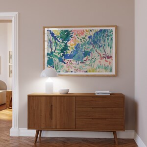 Pode incluir: Um aparador de madeira com duas portas e tr&ecirc;s gavetas. O aparador &eacute; de estilo moderno de meados do s&eacute;culo com pernas afiladas. Uma l&acirc;mpada branca e uma tigela branca est&atilde;o em cima do aparador. Uma pintura emoldurada de uma paisagem colorida est&aacute; na parede acima do aparador.