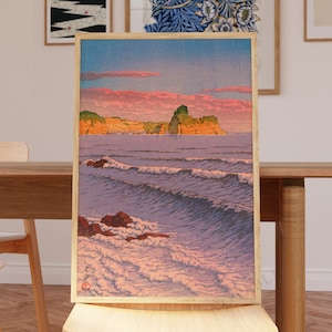 Könnte beinhalten: Ein japanischer Holzschnitt aus der Zeit, der eine Meereslandschaft mit einer kleinen Insel in der Ferne zeigt. Der Himmel ist in sanftem Rosa und Orange gehalten, die Wellen sind tiefblau. Der Druck ist in einem einfachen Holzrahmen gerahmt.