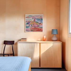 May include: A light blue bedspread with a wooden nightstand with two doors. A framed painting of a colorful landscape hangs on the wall above the nightstand. A lamp with a blue lampshade sits on the nightstand.