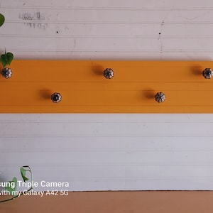 May include: A yellow wooden shelf with five decorative knobs. The knobs are black and white with a floral pattern. The shelf is mounted on a white wall.