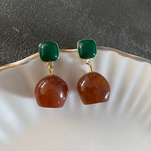 May include: A pair of earrings featuring a square, dark green top and a rounded, amber-colored bottom. The earrings are suspended by a gold-colored wire and are displayed on a white, shell-shaped dish.