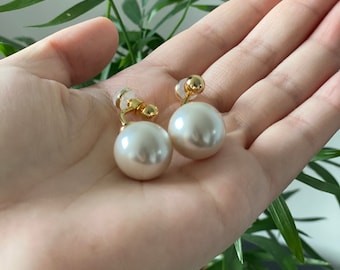 May include: A pair of clip-on earrings featuring large, round, faux pearls. The earrings have a gold-colored metal setting and clip-on backs. The pearls are a creamy white color and have a smooth, glossy finish.