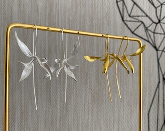 May include: Two pairs of floral earrings, one silver and one gold, are displayed on a gold stand. The earrings have intricate, petal-like designs with long, slender drops. The background features a textured grey wall and a geometric artwork.