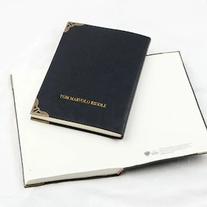 Peut inclure: Un journal en cuir noir avec des protège-coins dorés et le nom "TOM MARVOLO RIDDLE" en lettres dorées. Le journal est ouvert pour révéler des pages blanches, avec un second journal ouvert en dessous.