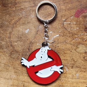 Peut inclure: Un porte-clés argenté avec un logo Ghostbusters rouge et blanc. Le logo représente un fantôme blanc avec un symbole rouge de non.