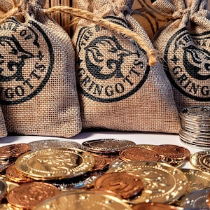 May include: Five tan burlap drawstring pouches each printed with a black circular logo reading The Bank of Gringotts and a dragon illustration. Below the bags are scattered piles of gold, silver, and copper-colored metal coins.