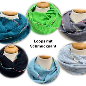 May include: Circular scarves in various colors, including teal, green, gray, black, and light blue, are displayed on mannequin heads. Each scarf features a decorative, colorful trim. The text "Loops mit Schmucknaht" is visible.