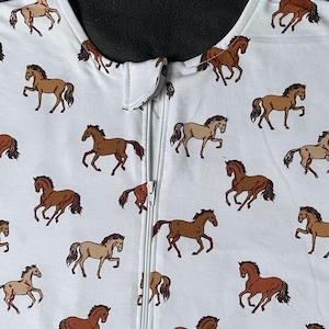 May include: White baby sleep sack featuring a repeating pattern of brown horses. The sleep sack has a full-length zip down the front and a rounded neckline. The horses are in various poses.