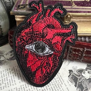 Può includere: Toppa ricamata di un cuore anatomico rosso con contorno nero e un solo occhio bianco al centro.