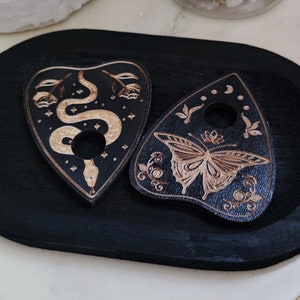 May include: Two black wooden planchette boards with gold designs. One planchette features a snake with a star pattern and the other features a butterfly with floral designs.