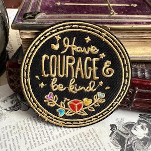 Könnte beinhalten: Ein schwarzer gestickter Aufnäher mit goldfarbenem Garn. Der Aufnäher zeigt ein florales Design mit einer roten Blume und dem Text "Have Courage & be kind."