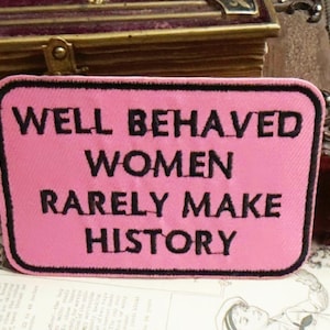 Könnte beinhalten: Ein rosa rechteckiger Aufnäher mit schwarzem Text, der lautet: "WELL BEHAVED WOMEN RARELY MAKE HISTORY". Der Aufnäher hat einen schwarzen Rand und befindet sich vor einem Hintergrund aus Vintage-Büchern und Papier.