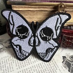 Könnte beinhalten: Ein weißer gestickter Patch mit einer Schmetterlingsform, der schwarze schädelartige Augen und ein detailliertes Design aufweist. Der Schmetterling ist ein beliebtes Motiv in der Gothic- und Dark-Mode.