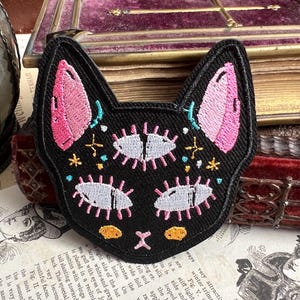 Puede incluir: Parche de hierro negro bordado de un gato con orejas rosas, ojos blancos y detalles amarillos. Los ojos del gato están rodeados de estrellas y otros pequeños detalles.