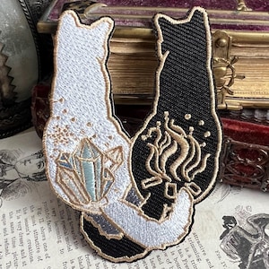 Peut inclure: Deux patchs thermocollants brodés de chats noirs et blancs. Le chat blanc tient un cristal bleu et le chat noir tient une flamme dorée.