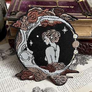 Op de afbeelding: Geborduurde strijkpatch met een vrouw die voor een halve maan met sterren staat. De maan is wit en de vrouw is wit met bruin haar. De achtergrond is zwart met bruine wolken.