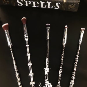 Könnte beinhalten: Sechs silberne Make-up-Pinsel mit schwarzen Griffen in Form von Zauberstäben. Die Pinsel haben schwarze Borsten und sind in einer Reihe auf einer schwarzen Oberfläche angeordnet. Die Pinsel sind mit "POTIONS SPELLS" beschriftet.
