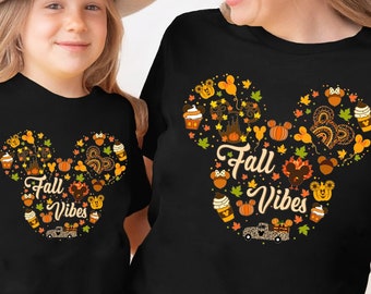 May include: Black t-shirts with a fall-themed design in the shape of Mickey Mouse's head. The design includes pumpkins, leaves, and the text "Fall Vibes". A mother and child are wearing the shirts.