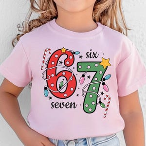 May include: A light pink t-shirt with the numbers "6" and "7" in a Christmas theme. The number "6" is red with candy cane accents, and the number "7" is green with a yellow star. The word "six" and "seven" are also on the shirt.