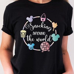 May include: Black t-shirt featuring a colorful design with the text "Snacking Around the World" in a circular arrangement. The design includes icons of snacks and Mickey Mouse silhouettes.