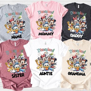 May include: Assorted-color Disneyland 2026 graphic t-shirts. The shirts feature a cartoon image of Mickey Mouse, Minnie Mouse, Donald Duck, and other Disney characters. Text on the shirts includes "Name", "Mommy", "Daddy", "Sister", "Auntie", and "Grandma".