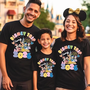 K&ouml;nnte beinhalten: Drei Personen tragen passende schwarze T-Shirts mit einer farbenfrohen Disney-Grafik. Die Shirts tragen die Aufschrift "Disney Trip" mit Platz f&uuml;r einen Namen und dem Jahr "2026". Die Grafik zeigt Micky Maus, Minnie Maus und andere Disney-Figuren.