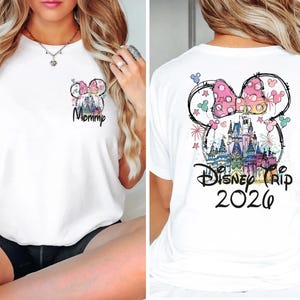 May include: White t-shirt featuring a colorful Minnie Mouse design with a castle and fireworks. The front of the shirt has the word "Mommy" in script. The back of the shirt says "Disney Trip 2026".