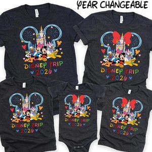 May include: A collection of dark gray t-shirts and a onesie featuring a colorful Disney design. The design includes Mickey Mouse ears, a castle, and various Disney characters. The text "DISNEY TRIP 2026" is displayed in rainbow colors.