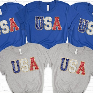 May include: Four t-shirts with the word "USA" in red, white, and blue sequins. The shirts are on a white brick background.
