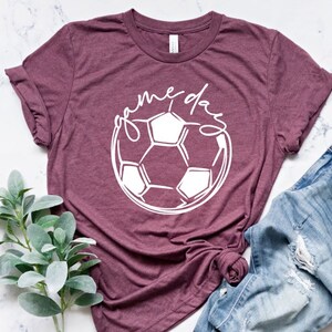 May include: A maroon t-shirt with a white soccer ball graphic and the text "game day" in a cursive font.