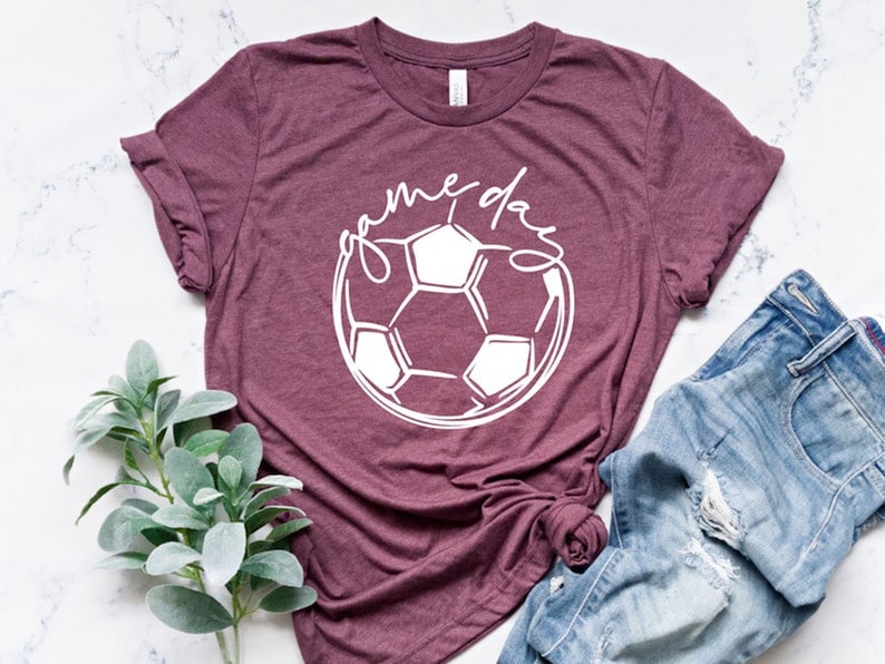 May include: A maroon t-shirt with a white soccer ball graphic and the text "game day" in a cursive font.