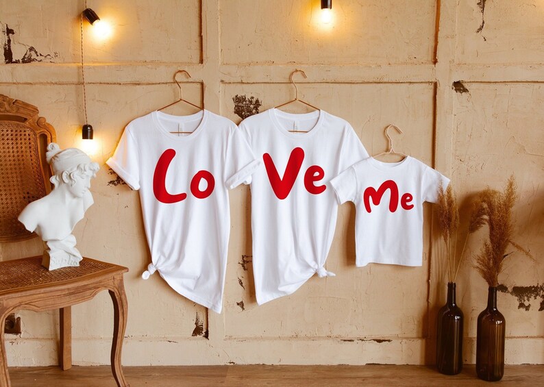 Puede incluir: Tres camisetas blancas con letras rojas que deletrean "Love Me". Las camisetas est&aacute;n colgadas en perchas contra una pared de color marr&oacute;n claro.