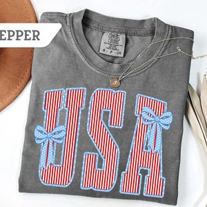 Może przedstawiać: Szara koszulka Comfort Colors z napisem "USA" w czerwono-białe paski i niebieskie kokardki w kropki. Srebrny naszyjnik z wisiorkiem spoczywa na koszulce. Koszulka jest złożona na białej powierzchni.