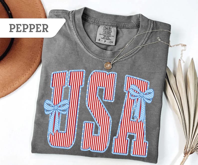 Może przedstawiać: Szara koszulka Comfort Colors z napisem "USA" w czerwono-białe paski i niebieskie kokardki w kropki. Srebrny naszyjnik z wisiorkiem spoczywa na koszulce. Koszulka jest złożona na białej powierzchni.