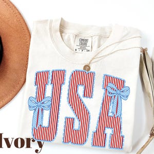 Może przedstawiać: Kremowa koszulka z napisem "USA" w czerwono-białe paski, obramowanym na niebiesko. Jasnoniebieskie kokardy z białymi kropkami zdobią litery. Na zdjęciu widoczny jest r&oacute;wnież złoty naszyjnik i brązowy kapelusz.