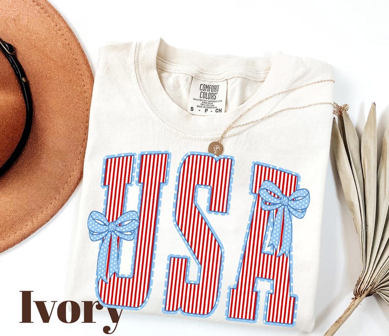 Może przedstawiać: Kremowa koszulka z napisem "USA" w czerwono-białe paski, obramowanym na niebiesko. Jasnoniebieskie kokardy z białymi kropkami zdobią litery. Na zdjęciu widoczny jest r&oacute;wnież złoty naszyjnik i brązowy kapelusz.
