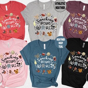 May include: Several short-sleeved t-shirts in shades like red, grey, and black. Each shirt displays the phrase "Snacking Around the World" in a circular design, accompanied by food and character illustrations. The shirts are available in multiple colours.