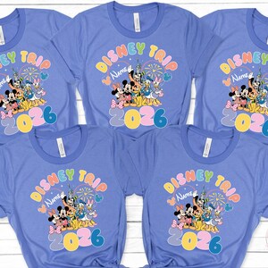 K&ouml;nnte beinhalten: Mehrere hellblaue T-Shirts mit einem farbenfrohen Disney Trip-Design. Das Design zeigt Micky und Minnie Maus, Donald und Daisy Duck sowie den Text "Disney Trip Name 2026".