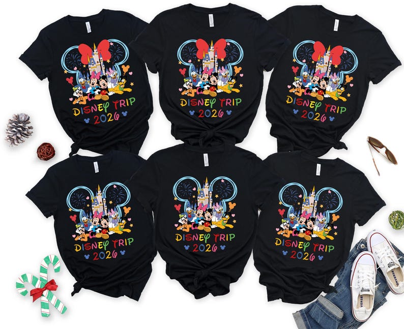 May include: Six black t-shirts with a colourful Disney graphic featuring Mickey Mouse, Minnie Mouse, and other characters in front of a castle. The text "DISNEY TRIP 2026" is printed below the graphic in rainbow colours. The shirts are displayed with holiday decorations and a pair of jeans.