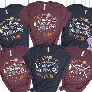 May include: Several t-shirts in heather navy and maroon, each with a circular design reading "Snacking Around the World." The design incorporates snack illustrations and the Disney World logo. Available in various colours, these t-shirts are ideal for theme park fans.