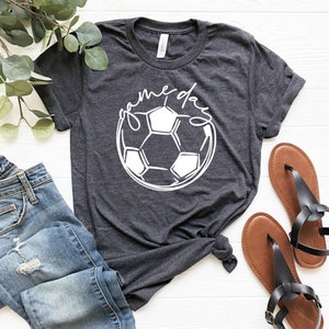 May include: A dark grey t-shirt with a white soccer ball graphic and the text "game day" in a cursive font.