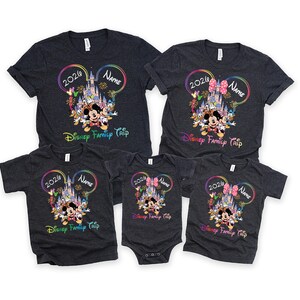 May include: A set of dark grey t-shirts and a onesie with a colourful Disney design. The design includes Mickey Mouse, a castle, and the text "2026", "Name", and "Disney Family Trip". The shirts are short-sleeved.