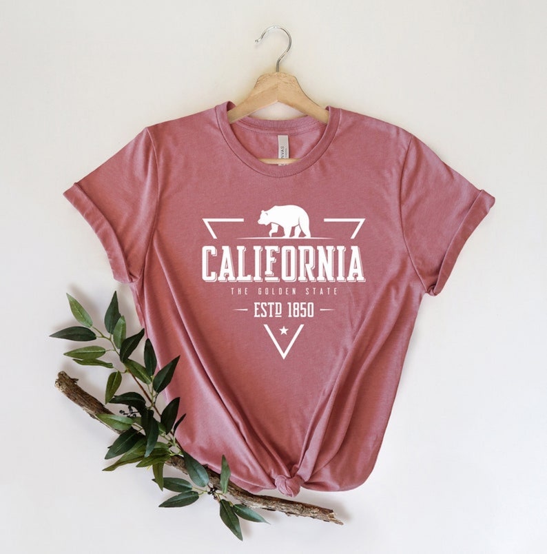 May include: A dusty rose t-shirt with a white graphic of a bear inside a triangle. The graphic says "California, The Golden State, Est. 1850".