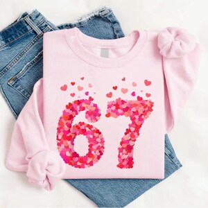 May include: A light pink sweatshirt with the number "67" in a heart-shaped design. The number is composed of various shades of pink and red hearts. The sweatshirt is paired with blue jeans and a matching scrunchie.