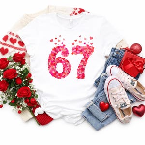 May include: White t-shirt with the number 67 made of pink and red hearts. The shirt is displayed with a bouquet of red roses, a red gift box, and a pair of light-colored sneakers on a pair of blue jeans. A red heart ornament is also present.
