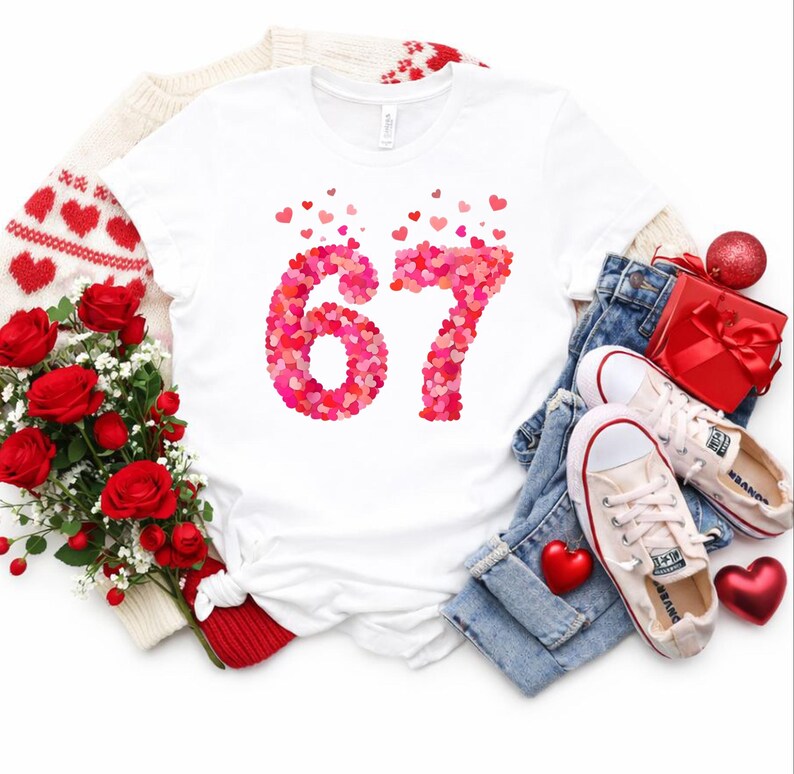 May include: White t-shirt with the number 67 made of pink and red hearts. The shirt is displayed with a bouquet of red roses, a red gift box, and a pair of light-colored sneakers on a pair of blue jeans. A red heart ornament is also present.